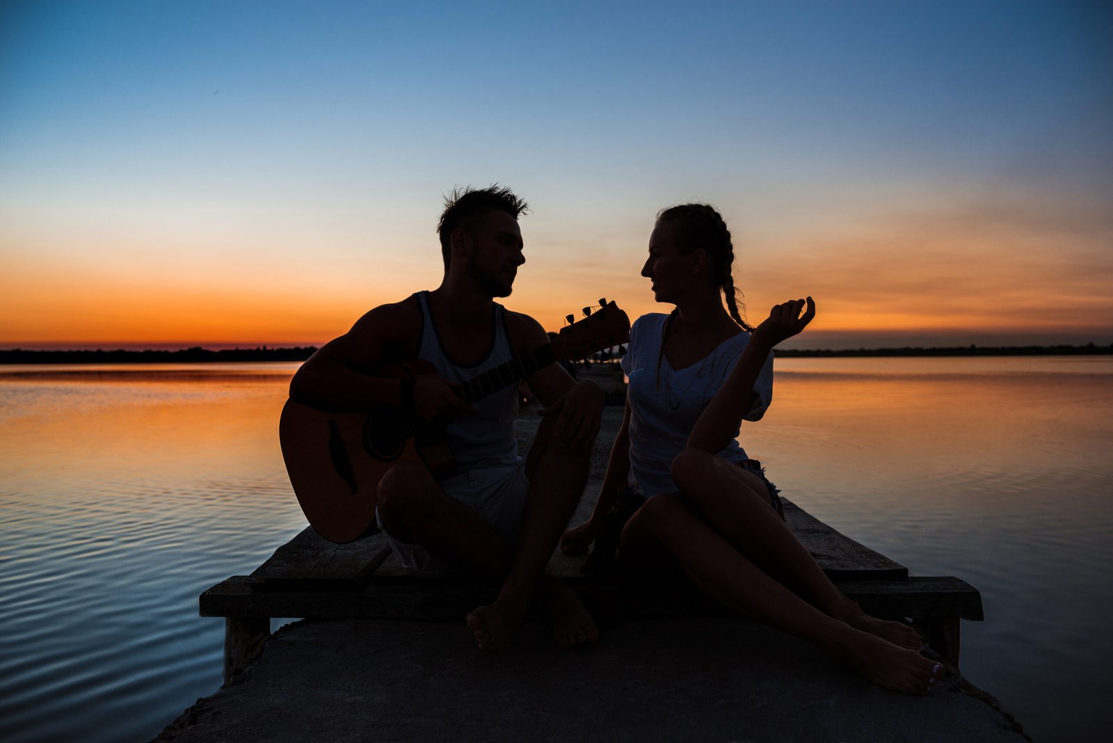 Silhouettes of young beautiful couple resting rejoicing at sunrise near lake.
